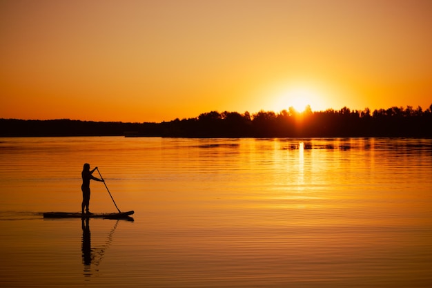 Silhueta de mulher embarcando sozinho com remo nas mãos no lago calmo com o sol quase se pondo abaixo do horizonte no fundo cobrindo a superfície da água com cor laranja Estilo de vida ativo