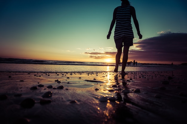 Silhueta de mulher na praia do pôr do sol, garota caminhando ao longo da costa do mar sob o sol de verão