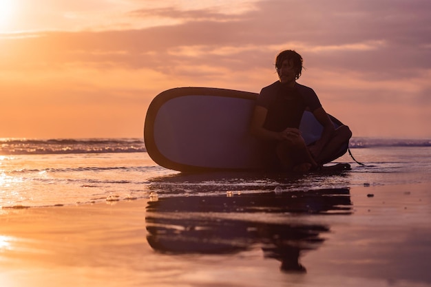 Silhueta de surf man sentado com uma prancha de surf na praia à beira-mar na hora do pôr do sol