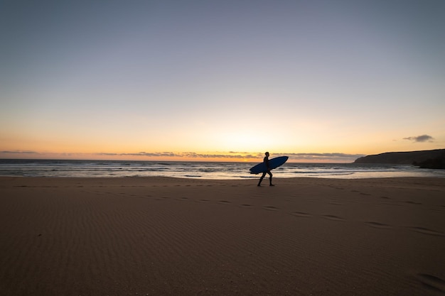 Silhueta de um homem adulto caminhando na praia vazia segurando uma prancha de surf