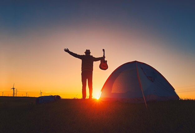 Foto silhueta de um homem com os braços estendidos segurando uma guitarra em uma tenda em um campo contra o céu durante o pôr do sol