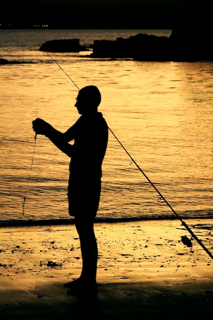 Foto silhueta de um homem com uma vara de pesca de pé contra o mar durante o pôr do sol