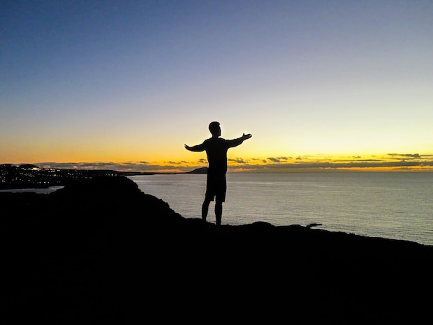 Silhueta de um homem de pé na costa contra o céu durante o pôr do sol