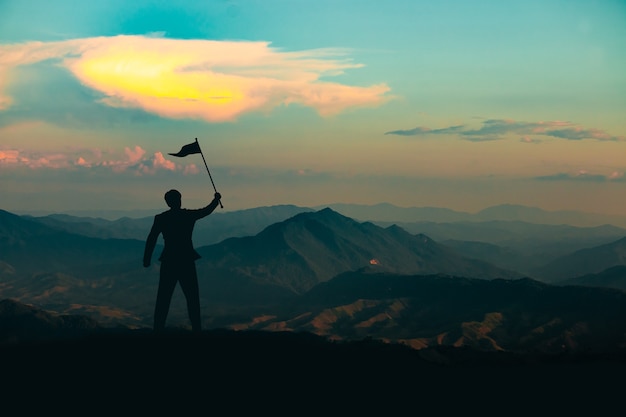 Silhueta de um homem em pé com uma bandeira da vitória no topo da montanha no fundo do céu ao nascer do sol, conceito de negócios, sucesso, liderança e realização
