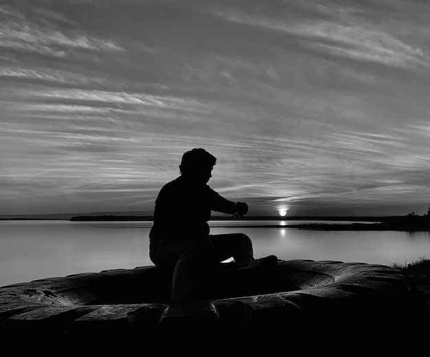 Foto silhueta de um homem sentado em uma rocha ao lado do mar contra o céu durante o pôr do sol