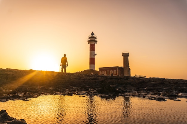 Silhueta de um jovem ao pôr do sol do farol de Toston, Punta Ballena, perto da cidade de El Cotillo, ilha de Fuerteventura, nas Ilhas Canárias. Espanha