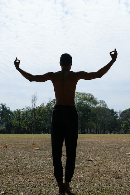 Silhueta de um jovem brasileiro sem camisa, de costas e braços abertos em um parque público.