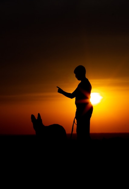 Foto silhueta de um jovem macho andando com um cachorro no campo ao pôr do sol, cara com um pastor alemão curtindo a natureza, menino sentado em uma laje de concreto perto de seu animal de estimação, o conceito de lazer ativo