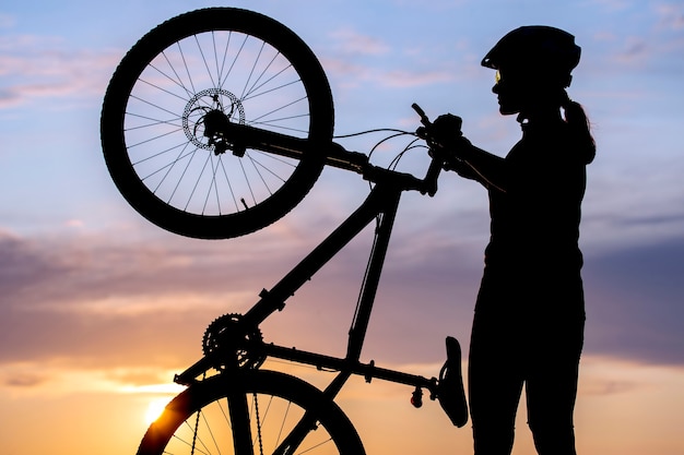 Silhueta de uma garota ciclista com uma bicicleta no fundo de um céu ao pôr do sol. Estilo de vida saudável e esporte. Lazer e hobbies