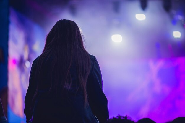 Silhueta de uma grande multidão em show contra um palco bem iluminado. Concerto de rock noturno com pessoas se divertindo, levantando as mãos no ar e aplaudindo os músicos.