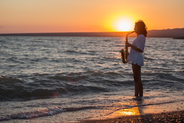 Silhueta de uma jovem mulher sexy tocando saxofone na praia ao pôr do sol