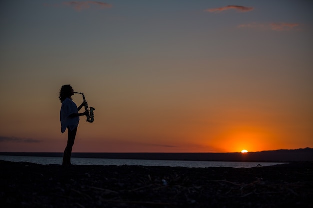 Silhueta de uma jovem mulher sexy tocando saxofone na praia ao pôr do sol