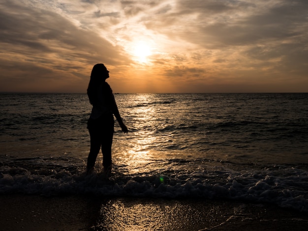 Foto silhueta de uma jovem na praia. jovem está caminhando ao pôr do sol à beira-mar. turista na praia durante as férias