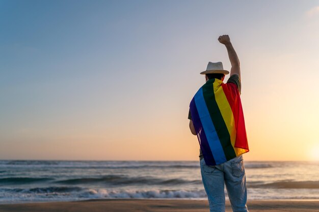 Silhueta de uma pessoa com bandeira arco-íris LGTBQ com a mão levantada na praia ao pôr do sol