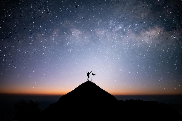 Silhueta do empresário de pé no topo da montanha sobre o céu noturno com estrela Via Láctea e luz solar com bandeira Ele levantou os dois braços mostrando alegria e sucesso