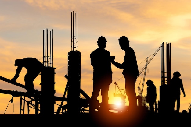 Silhueta do engenheiro e do trabalhador verificando o projeto no canteiro de obras de fundo do canteiro de obras ao pôr do sol à noite