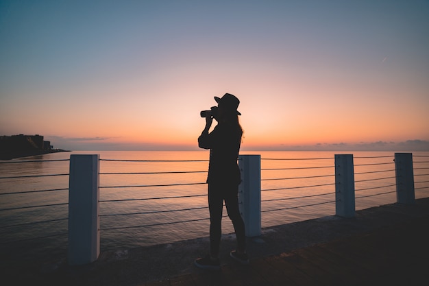 Silhueta do fotógrafo de mulher de chapéu com câmera digital durante a tomada de fotos do mar no cais ao pôr do sol