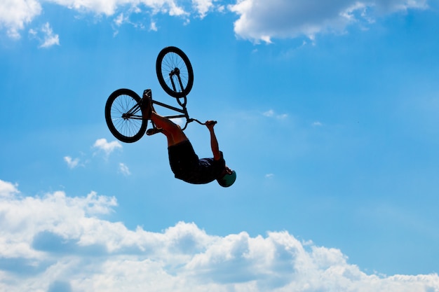 Silhueta do homem que salta na bicicleta contra um céu azul com nuvens brancas. Cara executa truques em uma bicicleta