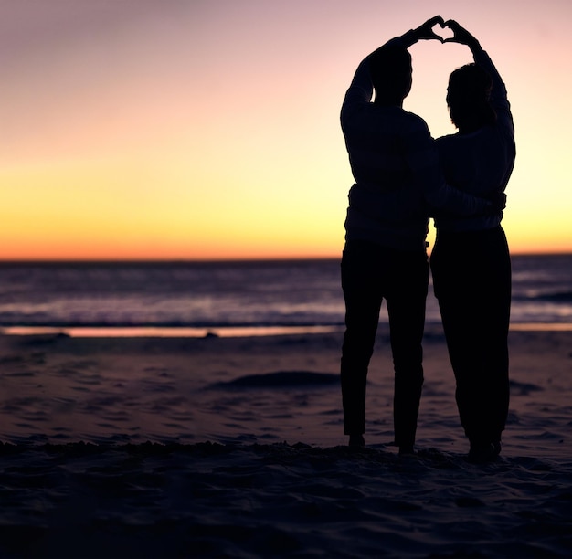 Silhueta do pôr do sol de casal e praia com mãos de sinal de coração se unindo e amando de férias para lua de mel Homem mulher e sinal de mão romântico pelo oceano com sol do entardecer para romance juntos na natureza