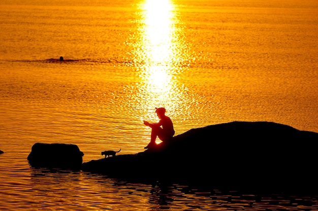 Silhueta do sol do homem sentado em um lago. Luz solar laranja ao ar livre do homem descansando.