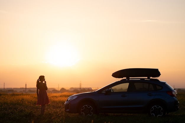 Silhueta escura de mulher solitária relaxante perto de seu carro em Prado gramado, apreciando a vista do colorido do nascer do sol. Jovem motorista do sexo feminino descansando durante a viagem ao lado do veículo SUV.