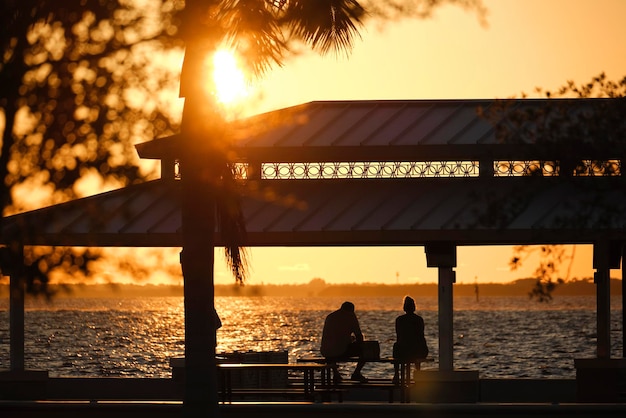 Silhueta escura de pessoas descansando sob o telhado de alcova na costa do mar em parque público ao pôr do sol