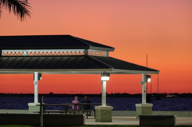 Silhueta escura de pessoas descansando sob o telhado de alcova na costa do mar em parque público ao pôr do sol