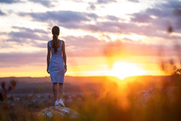 Silhueta escura de uma jovem mulher com vestido de verão em pé ao ar livre, apreciando a vista da natureza ao pôr do sol.