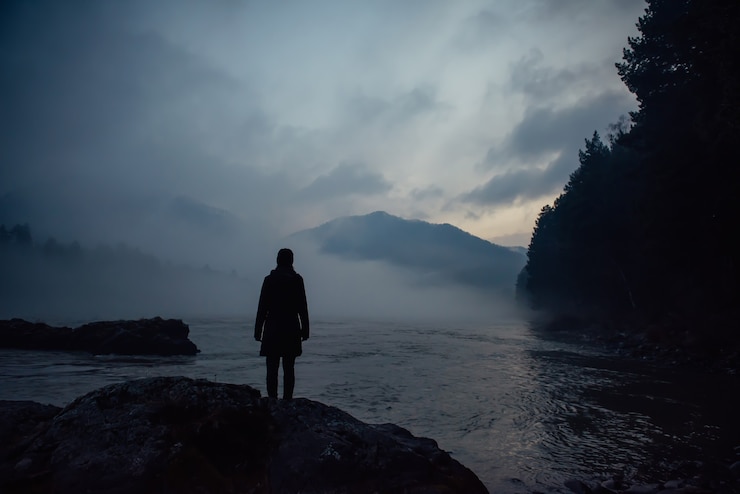 Foto silhueta humana sozinha na névoa branca contra as montanhas e o rio. nevoeiro espesso no crepúsculo da noite. atmosfera misteriosa. reflexão, meditação.