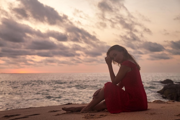 Silhueta linda mulher de vestido vermelho sentado na praia do oceano ao fundo do sol Senhora na praia do mar nas férias de verão