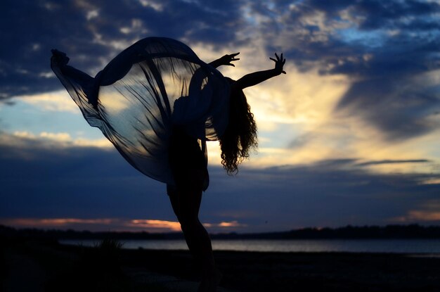 Silhueta de una mujer bailando en la orilla del mar contra el cielo al anochecer