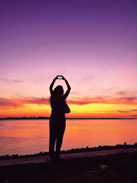 Foto silhueta de mujer gestando en forma de corazón contra el mar durante la puesta de sol