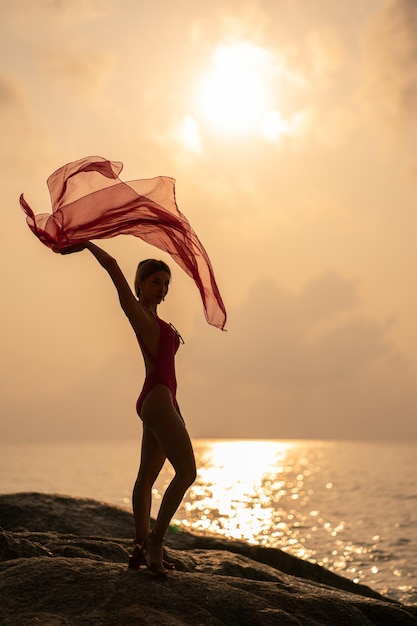 silhueta: Mulher vestindo maiô rosa de uma peça segurando lenço no vento desfrutar do momento romântico do pôr do sol na pedra, praia tropical.