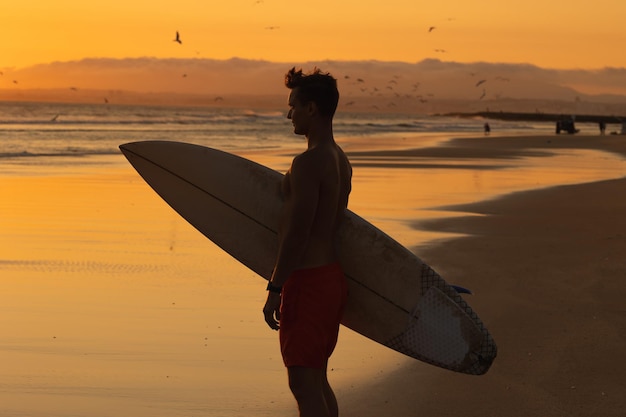 Silhueta negra de um homem atlético em pé na praia segurando uma prancha de surf ao pôr do sol