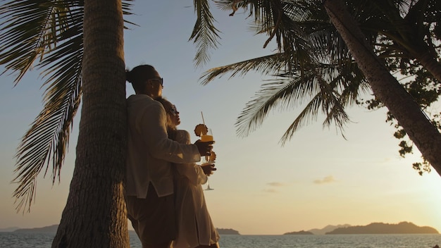 SILHUETA OURO ESCURO SUNSET VIEW Casal apaixonado na camisa branca e óculos de sol perto de palmeira beijando abraçando relaxando e bebendo coquetel Conceito férias de verão tropicais férias amor de casamento