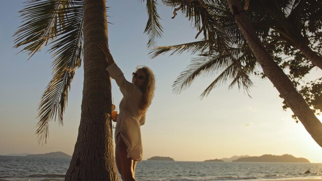 SILHUETA OURO ESCURO SUNSET VIEW Mulher bonita bebendo coquetel de abacaxi Pina Colada perto de palmeira Mulher em óculos de sol de camisa branca Conceito descanso mar tropical viajando turismo férias de verão