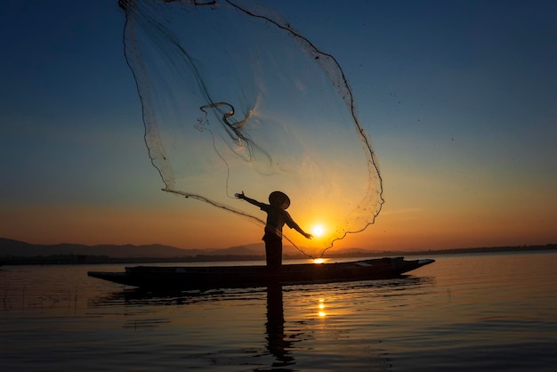 Silhueta de un pescador arrojando una red de pesca en el mar contra el cielo durante el amanecer