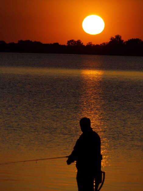 Foto silhueta de un pescador de pie frente al lago durante la puesta de sol