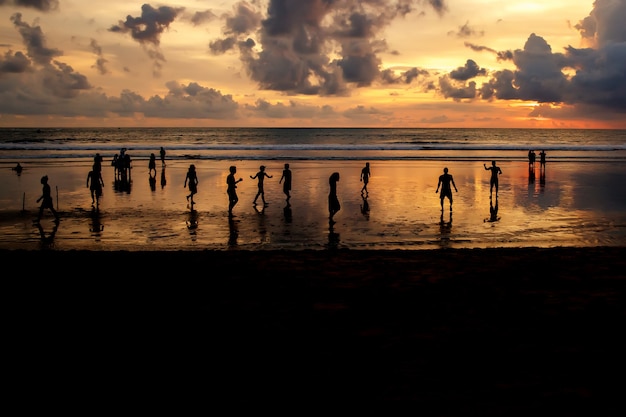 silhuetas de caras que jogam futebol ao pôr do sol na praia do oceano.