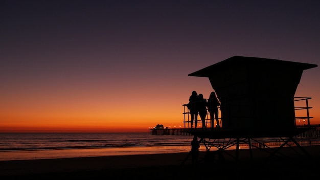 Silhuetas de meninas adolescentes perto da torre do salva-vidas, amigos na praia do Oceano Pacífico, pôr do sol em Oceanside, Califórnia, EUA