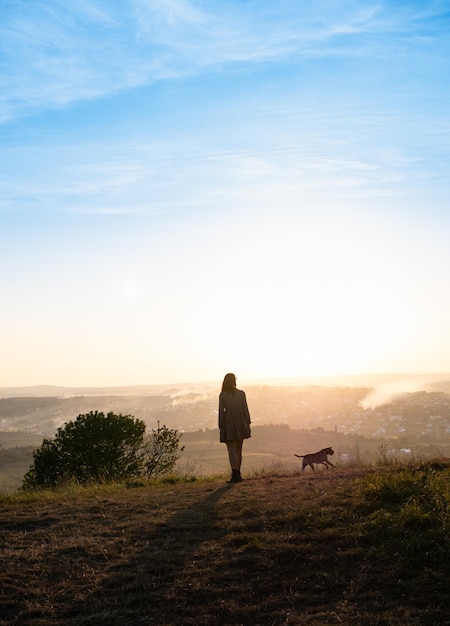 Silhuetas de mulher adorável brincando com seu cachorro fofo durante o pôr do sol