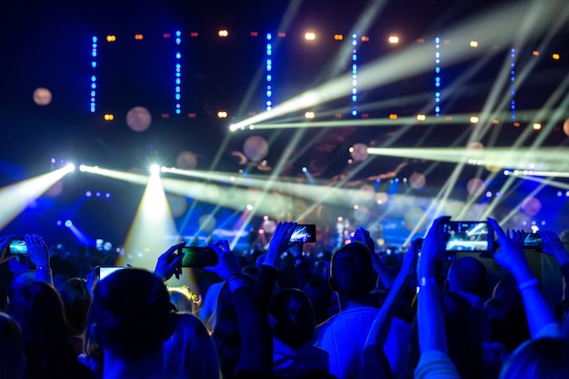 Silhuetas de multidões de espectadores em um show com smartphones nas mãos. A cena é lindamente iluminada por holofotes.