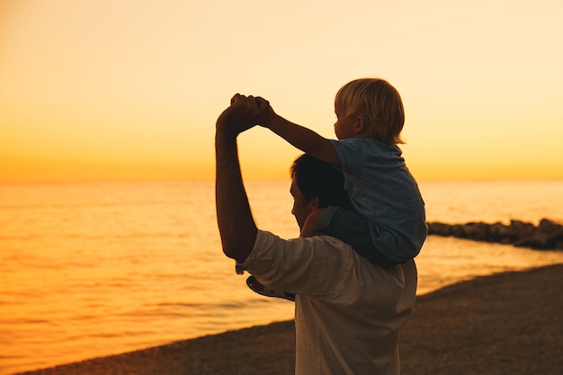 Silhuetas de pai e filho no céu pôr do sol Família amorosa e férias de verão Homem e criança menino brincando juntos ao ar livre em uma praia do mar Pai carregando criança nas costas com os braços levantados na natureza