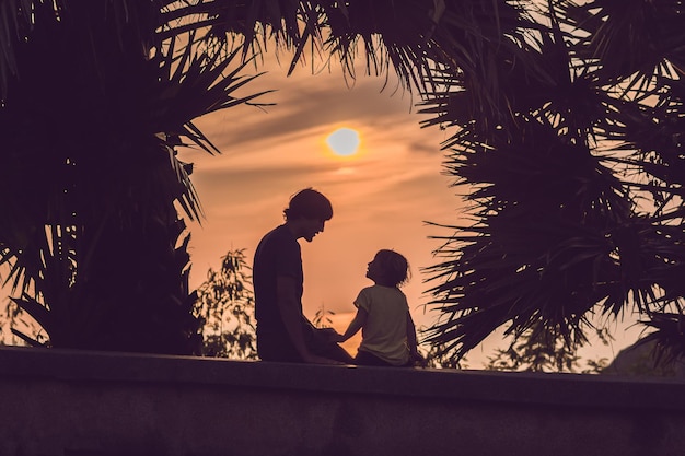 Silhuetas de pai e filho, que se encontram com o pôr do sol nos trópicos, tendo como pano de fundo palmeiras