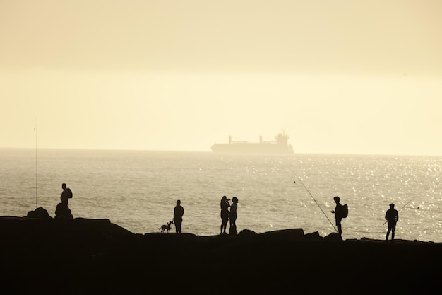 Silhuetas de pessoas em pé à beira-mar pescando e passando o tempo no pôr do sol nebuloso