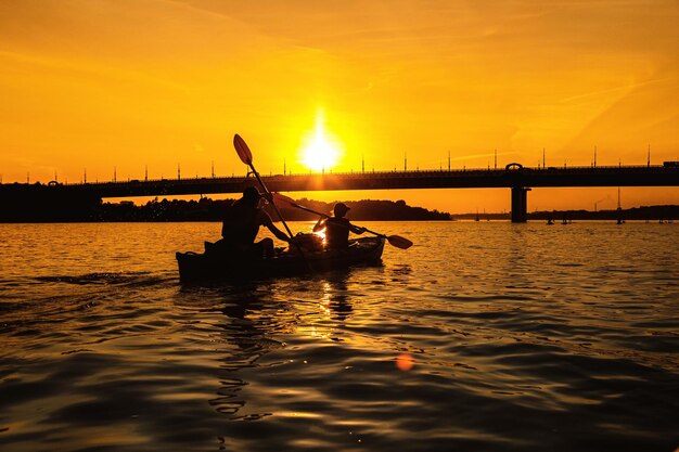 Silhuetas de pessoas em um caiaque ao pôr do sol. Casal jovem está passeando de barco no rio