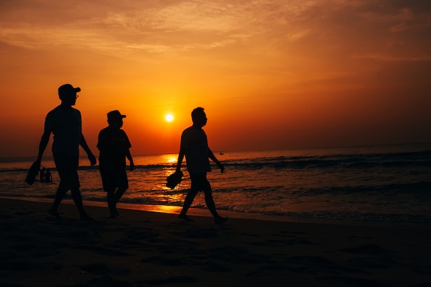 Silhuetas de três homens caminhando na praia ao pôr do sol