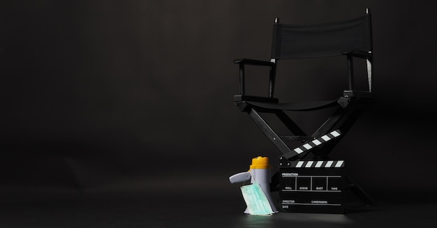 Silla de director NEGRA con claqueta amarilla o tablero de chapaleta de película y máscara facial, megáfono sobre fondo negro.Se usa en producción de video o industria cinematográfica y cinematográfica.