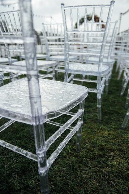 Sillas de cristal blanco sobre la hierba verde. Arco de medio punto para una boda en el fondo. Día lluvioso