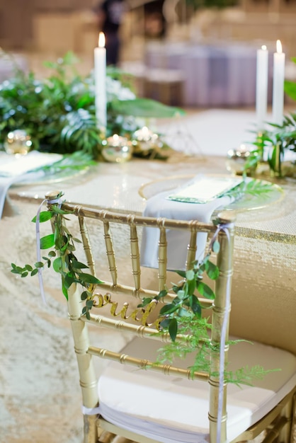 Sillas doradas con letrero para la novia y el novio con ramitas verdes de ruscus Salón de eventos con decoraciones doradas y hojas de palmeras tropicales